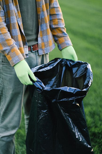 A person wearing a checked shirt over a green T-shirt and light grey trousers is standing outdoors on a grassy area, holding open a large black waste bag lined with a reflective inner layer. The individual is wearing light green gloves and appears to be preparing to dispose of rubbish into the bag, which is partially filled. The background shows a lush, green lawn, suggesting a park or garden setting. This scene reflects a process of waste collection or clearing, typical of private rubbish removal tasks undertaken by services such as those offered by companies specialising in independent rubbish clearance solutions. The environment is well-lit, with natural daylight illuminating the scene, emphasizing the practical aspect of rubbish handling and environmental cleanliness.