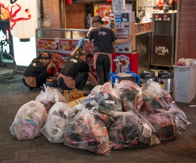 Several large transparent plastic bags filled with mixed rubbish, including clothing and miscellaneous waste, are piled on a paved sidewalk in front of a shop or fast-food outlet at night. In the background, three workers dressed in black uniforms with brown accents and wearing caps are actively engaged in clearing or handling additional waste near a red wall with a food counter and a microwave oven. To the right, there are two black refuse bins with lids and a trash container lined with black plastic. The scene is illuminated by artificial lighting, highlighting the textures of the crumpled plastic bags and the surrounding urban environment, suggesting an example of private on-site rubbish collection by a waste management service, and supporting the context of efficient rubbish removal in a commercial area.