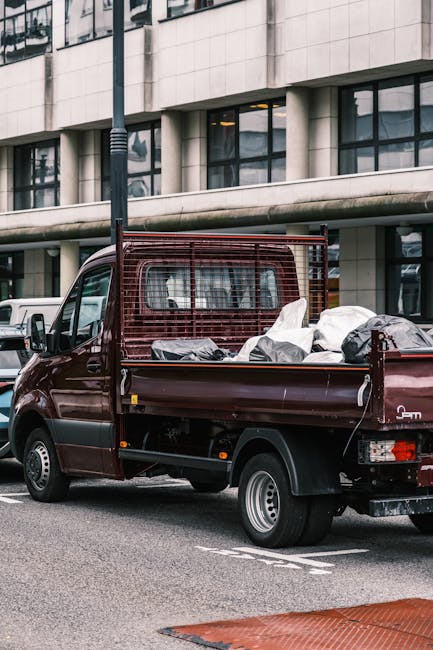 A small, dark red flatbed truck with an open cargo area is parked on an urban street in front of a modern, beige-colored office building with large, reflective glass windows. The truck's rear bed is loaded with various discarded items, including white and black bags, some of which appear to contain household waste or rubbish. A metal cage-like partition separates the cab from the load, and the items are haphazardly arranged, suggesting a private waste collection or rubbish clearance operation. The scene is well-lit, with natural daylight casting minimal shadows, emphasizing the textures of the bags and the truck's surface. The background shows additional parked vehicles and a lamppost, providing context for a busy city environment where independent rubbish removal services, such as those provided by [COMPANY_NAME], facilitate on-site clearance outside of local authority waste collection. This image illustrates the process of private rubbish disposal, often undertaken for quick and efficient clearance of household or commercial waste in an urban setting.