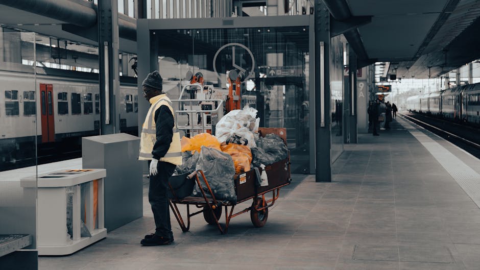 A narrow urban alleyway cluttered with large black and white garbage bags and loose rubbish, including cardboard boxes and assorted waste, piled up against a backdrop of makeshift cardboard stacks and debris. The scene is situated between tall, weathered buildings with graffiti on the walls, some metal grates over windows, and cables running along the facades. In the foreground, a wheeled rubbish bin covered with a worn, grey protective bag is positioned on the asphalt surface, which appears stained and uneven. The alley is dimly lit, with natural light filtering in from the open end, highlighting the clutter and the surrounding environment. This scene reflects a situation where private waste collection through an independent rubbish removal service, such as that offered by rubbishclearanceshepherdsbush.com, might be employed for on-site clearance of bulk waste and debris from urban areas.