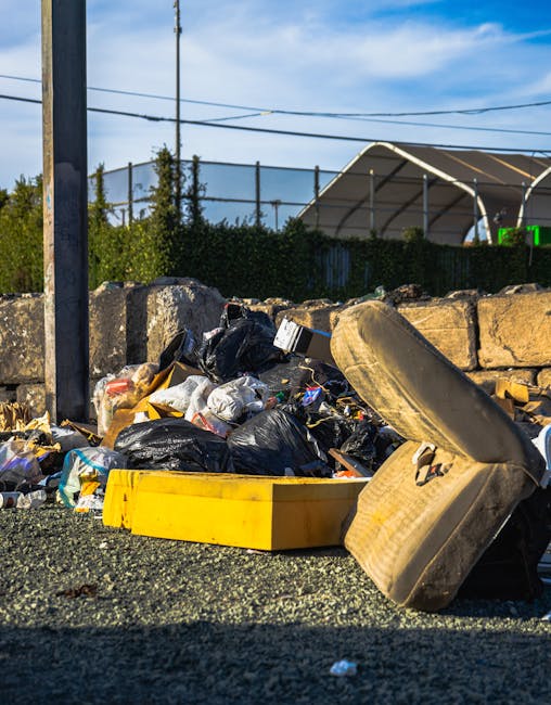 A close-up view of a collection of household rubbish and waste materials piled on a gravel surface outdoors. The waste includes black plastic garbage bags, a yellow plastic storage container, and an old, beige upholstered armchair with visible fabric wear and some staining, lying on its side. Behind the pile, a low stone wall constructed from rough-hewn stones separates the area from a background featuring green hedge fencing, utility poles with black wires, and a partially visible large white tent or canopy structure with a curved roof. The scene is lit by natural daylight, creating shadows cast by the objects, and the environment appears to be an informal dumping area or a site set for waste collection, demonstrating a typical example of private rubbish disposal that might be handled by independent waste removal services like those offered by Shepherds Bush rubbish removal prices explained W12. The overall setting reflects a functional outdoor space used temporarily for waste storage or debris removal, aligned with rubbish clearance activities in the local area.